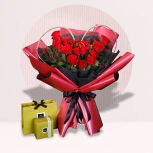 A bouquet of red roses is arranged in a heart shape and wrapped in red and black paper. A golden bow and a golden gift box with a gold ribbon are placed next to the bouquet.
