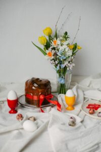 A glazed Easter cake decorated with a red ribbon, surrounded by colorful eggs and a vase of tulips and daffodils, on a white tablecloth.