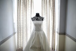 A white wedding dress with lace detailing hangs on a mannequin in front of a window.