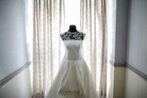A white wedding dress with lace detailing hangs on a mannequin in front of a window.