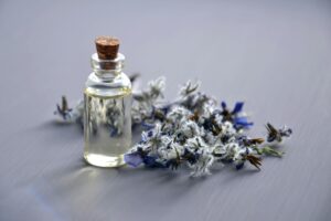 Small glass bottle of lavender oil with a cork stopper, next to a pile of dried lavender flowers on a gray background.