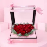 Heart-shaped box filled with red roses, greenery, and accented with black wrap and red ribbons, symbolizing love and devotion.