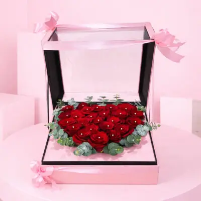 Heart-shaped box filled with red roses, greenery, and accented with black wrap and red ribbons, symbolizing love and devotion.