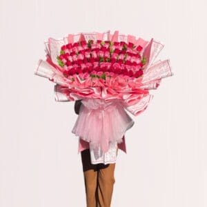 A large, opulent bouquet of pink and red roses is held by a person against a light, off-white background.