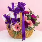 Treat and flower basket with pink and purple gerberas, white fillers, and purple ribbon decorations.