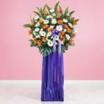 Flower stand with orange and white gerberas, greenery, and a purple ribboned base.