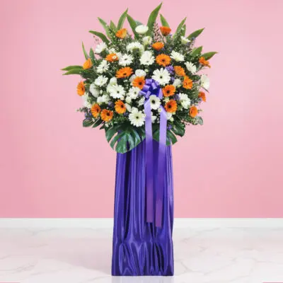 Flower stand with orange and white gerberas, greenery, and a purple ribboned base.