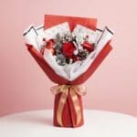 Christmas hand bouquet with red roses, berries, frosted leaves, silver ornaments in red-and-white wrapping.