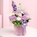 Lavender rose hatbox flower arrangement with pink roses, white flowers, greenery, and ribbon on a wooden table.