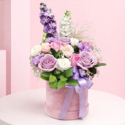 Lavender rose hatbox flower arrangement with pink roses, white flowers, greenery, and ribbon on a wooden table.
