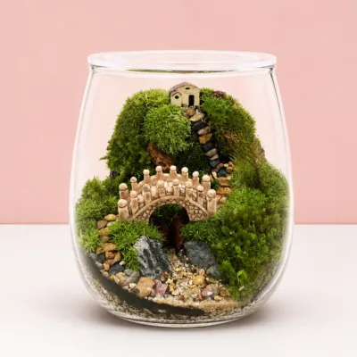 Enchanted Bridge Glass Bowl Terrarium featuring a miniature bridge, lush green moss, and a tiny cottage landscape arranged inside a decorative glass terrarium.