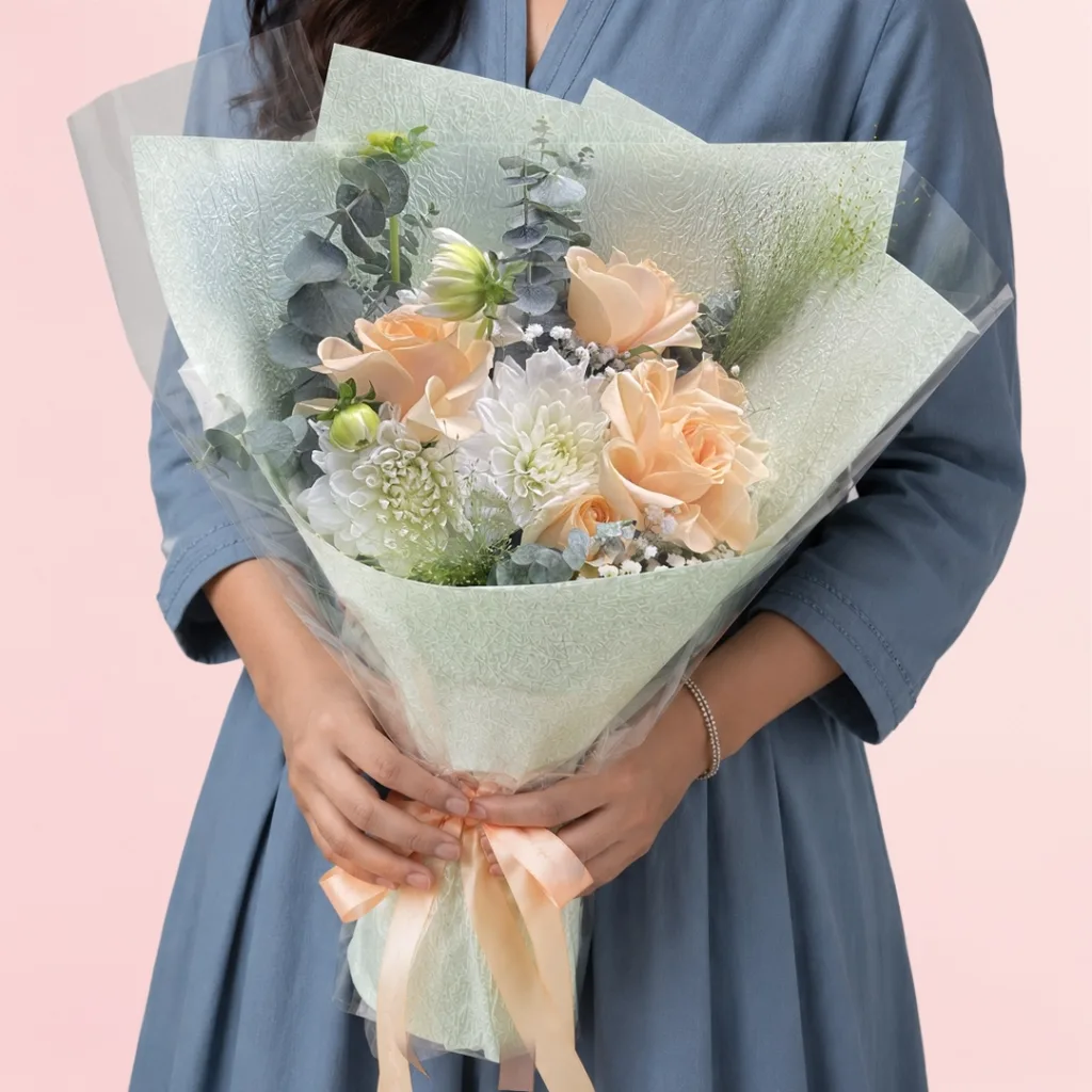 Fresh peach roses and chrysanthemum flowers are arranged in a white wrap with lush mixed greens and natural fillers.