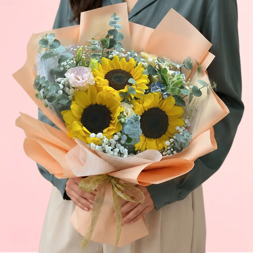 Three sunflowers with a pink carnation or garden rose, beautifully arranged with fresh green fillers wrapped in a pastel sheet.