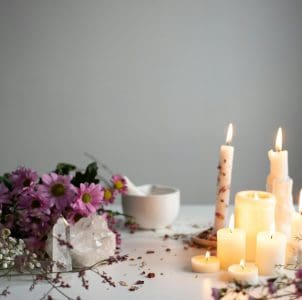 A collection of lit candles of varying sizes and shapes are arranged on a white surface. The candles cast a warm, yellow glow, with visible flames flickering atop each wick