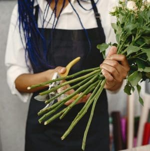 A person wearing a black apron and a white long-sleeved shirt is trimming the stems of a bunch of roses with yellow-handled pruning shears.